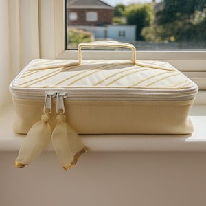 May include: A pale yellow cosmetic bag with a gold handle and zipper pulls. The top of the bag has gold striped detailing. Two cream-coloured tassels with gold trim hang from the zipper.