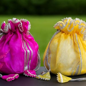 May include: Two drawstring pouches, one fuchsia and one yellow, sit side-by-side. Both have silver trim and drawstrings. The fuchsia pouch has silver stripes. The yellow pouch has a silver drawstring and trim. The background is a blurred green.