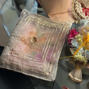 May include: A decorative, square, sheer cushion with a floral design. The cushion features a central flower with gold sequins and a gold border. The background is a soft pink and purple. Other decorative items are in the background.