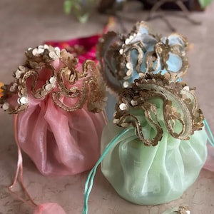 May include: Three pastel-coloured organza drawstring pouches: pink, light blue, and green. Each pouch features gold sequin trim and a matching drawstring. These pouches are likely for small gifts or favours.