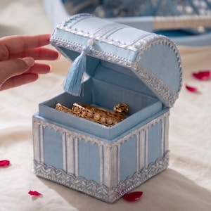 May include: A light blue fabric jewellery box with a hinged lid, featuring silver decorative trim, a matching light blue tassel, and a square base. The open box contains gold-toned bangles with circular accents inside the lined interior.