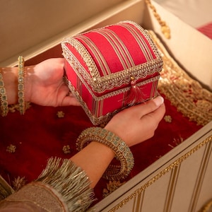 May include: A square red fabric box with a domed lid, featuring vertical gold stripes and intricate gold lace trim along the edges. The box includes a small red tassel hanging from the front centre, set within a larger cream-coloured chest.