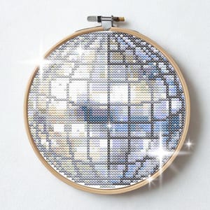 Puede incluir: Un bordado de punto de cruz en un aro de madera. El diseño presenta un patrón geométrico en tonos de azul, blanco y gris. El texto "Piesling stitch face" está en la parte inferior de la imagen.