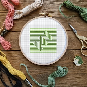 Puede incluir: Un bastidor de bordado circular de madera que contiene tela Aida blanca con un patrón de laberinto geométrico verde bordado en punto de cruz. Alrededor del bastidor hay madejas de hilo rosa, amarillo y verde, tijeras doradas y un alfiler de erizo.