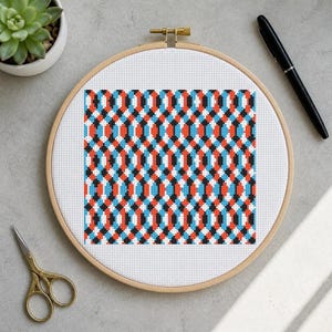 May include: A circular wooden embroidery hoop containing white Aida fabric with a geometric cross-stitch pattern. The design features repeating vertical columns of interlocking diamonds in vibrant shades of orange, bright blue, and black thread.