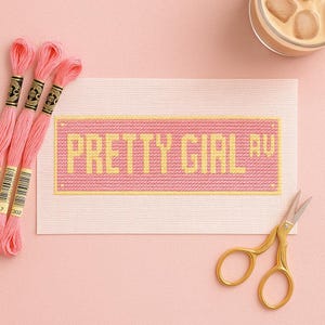 May include: A cross-stitch project with the words "PRETTY GIRL RU" in yellow on a pink background. Pink embroidery floss and gold scissors are also in the image. The project is on a light pink surface.