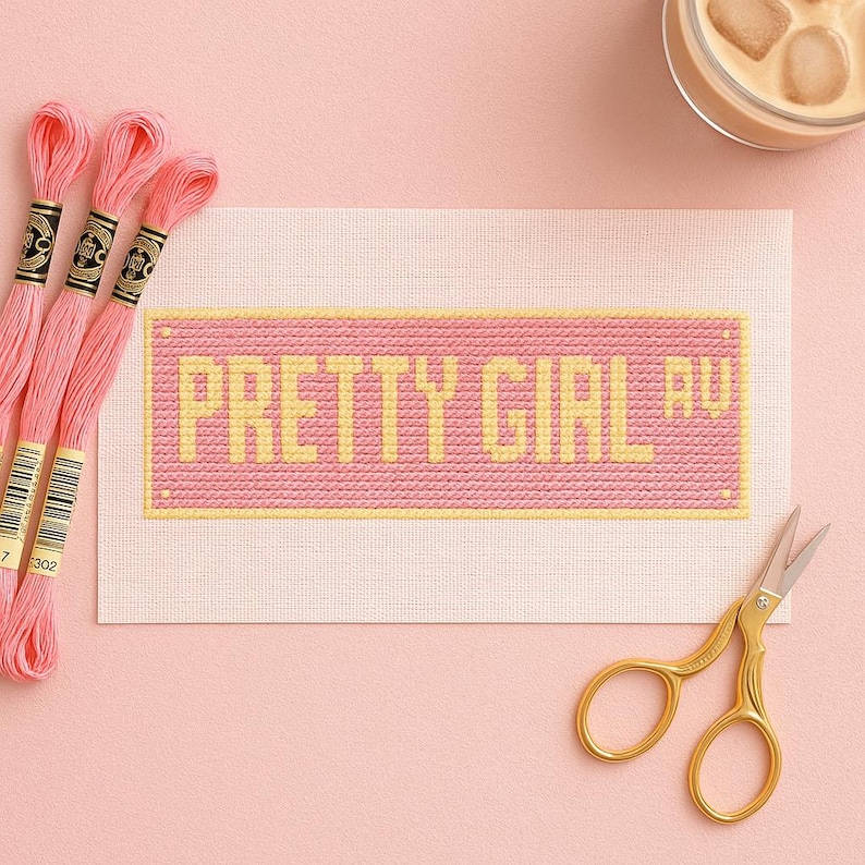 May include: A cross-stitch project with the words "PRETTY GIRL RU" in yellow on a pink background. Pink embroidery floss and gold scissors are also in the image. The project is on a light pink surface.