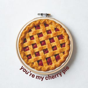 Puede incluir: Bordado de punto de cruz de una tarta de cerezas en un aro de madera. La tarta tiene una corteza de rejilla y un relleno rojo. El texto dice "you're my cherry pie" y "Piesling stitch face".