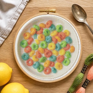 May include: A circular wooden embroidery hoop containing white fabric with a cross-stitch pattern of multicolored cereal loops in shades of red, orange, yellow, green, and blue. The hoop features a brass tension screw at the top center.