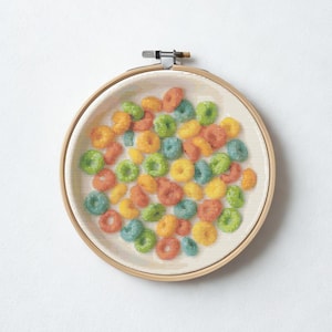 Op de afbeelding: Een borduurring met kruissteek met een kleurrijk ontbijtgranenontwerp. De ontbijtgranen zijn in verschillende kleuren, waaronder oranje, geel, groen en blauw. De tekst "Piesling stitch face" staat onderaan.