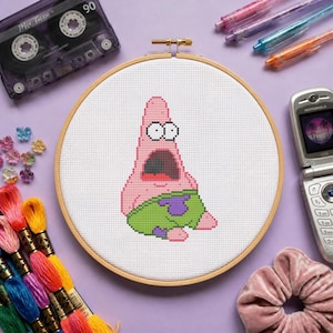 May include: A circular wooden embroidery hoop containing a white fabric panel with a cross-stitch design of a pink starfish character with wide eyes and an open mouth, wearing green shorts with purple flower patterns, centred on a light purple surface.