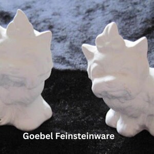 May include: Two white ceramic cat figurines, each in a seated position with a crown-like detail on their heads. The figurines are displayed against a dark background. The text "Goebel Feinsteinware" is visible at the bottom.