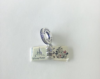 Könnte beinhalten: Ein silberner Charm-Anhänger in Form eines aufgeschlagenen Buches. Eine Seite zeigt eine Burg mit dem Text "Disney Parks", die andere eine Illustration von Mickey und Minnie. Der Charm hat einen Ring mit blauen Akzenten und dem Wort "Pandora".