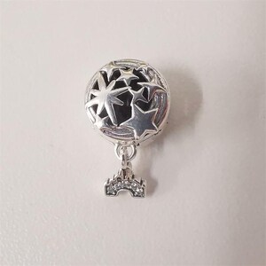 May include: A silver charm featuring a round design with star cut-outs. It has a dangling castle pendant with clear, sparkling details. This charm is likely for a bracelet or necklace.