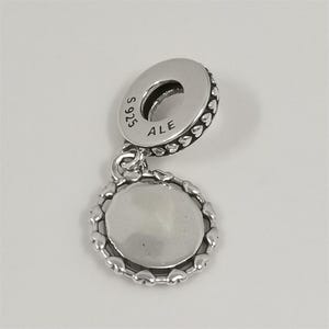 Puede incluir: Un charm de plata con un colgante redondo y un borde en forma de coraz&oacute;n. El charm tiene un lazo para sujetarlo a una pulsera o collar. La parte superior del charm tiene el texto "S 925 ALE".