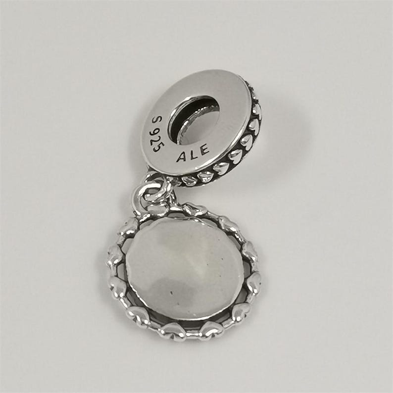 Puede incluir: Un charm de plata con un colgante redondo y un borde en forma de coraz&oacute;n. El charm tiene un lazo para sujetarlo a una pulsera o collar. La parte superior del charm tiene el texto "S 925 ALE".