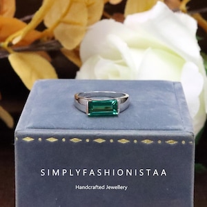 May include: A silver ring featuring a rectangular, emerald-green gemstone. The ring is presented on a blue velvet box, with the text "SIMPLYFASHIONISTAA" and "Handcrafted Jewellery". White and yellow flowers are in the background.