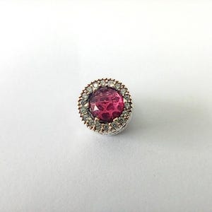 Puede incluir: Un charm circular con una piedra preciosa central redonda y facetada de color rosa intenso, rodeada por un halo de pequeñas piedras claras engastadas en un marco metálico. La pieza tiene una base plateada texturizada y un borde rosado.