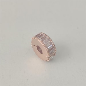 Op de afbeelding: Roségoudkleurige bedel met een ringvorm, versierd met rechthoekige, heldere kristallen. De bedel heeft een sluiting en is ontworpen om aan een armband of ketting te worden toegevoegd. De kristallen zijn rond de buitenrand geplaatst.