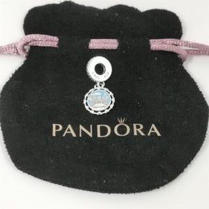 Puede incluir: Un charm de plata con un dise&ntilde;o azul claro y blanco, con un edificio, se muestra en una bolsa negra con cord&oacute;n. El charm est&aacute; marcado con "S925 ALE". La palabra "PANDORA" est&aacute; impresa en la bolsa.