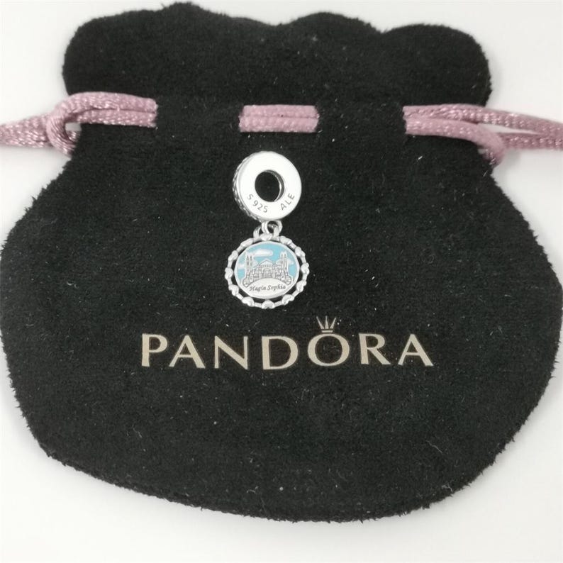 Puede incluir: Un charm de plata con un dise&ntilde;o azul claro y blanco, con un edificio, se muestra en una bolsa negra con cord&oacute;n. El charm est&aacute; marcado con "S925 ALE". La palabra "PANDORA" est&aacute; impresa en la bolsa.