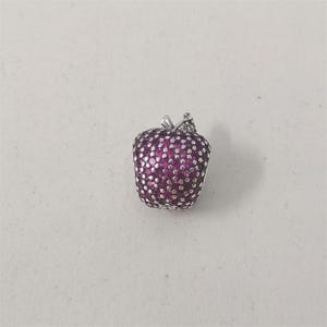 May include: A silver apple charm with a stem and leaf detail. The apple is covered in small, round, magenta-colored stones. The charm is likely for a bracelet or necklace.