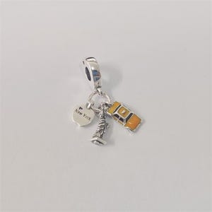 Può includere: Un charm d'argento con tre ciondoli: un'etichetta a forma di cuore con il testo "New York", una miniatura della Statua della Libertà e un taxi giallo con dettagli neri. Il charm è progettato per un braccialetto o una collana.