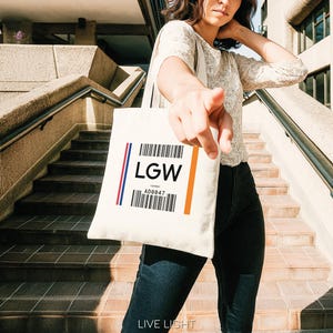 May include: A cream tote bag featuring the letters "LGW" and a barcode design. The bag has a red and blue stripe on the left and an orange stripe on the right. The bag is held by a person in front of a concrete staircase.