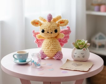 May include: A handmade crochet creature with yellow body, pink wings, and purple feet sits on a pink table. A small blue teacup, a jar with beads, and a cat-shaped planter with a succulent are also on the table. A handwritten note with a heart is visible.