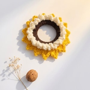 May include: A circular crochet hair accessory designed to resemble a sunflower, featuring a dark brown center, a ruffled cream-colored inner ring, and bright yellow pointed petals. A single whole walnut and a small dried flower sprig are nearby.