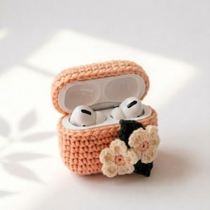May include: A peach-colored crochet case for wireless earbuds, featuring an open lid and two white earbuds inside. The exterior of the case is decorated with two cream-colored crocheted flowers with pink centers and dark green crocheted leaves.