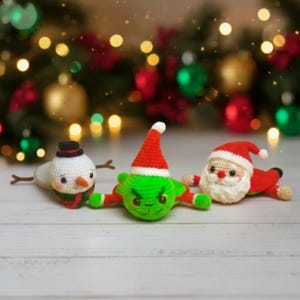 Puede incluir: Tres figuras navideñas de ganchillo: un muñeco de nieve con sombrero negro, un Grinch verde con gorro rojo y Papá Noel con traje y gorro rojos. Las figuras están tumbadas sobre una superficie de madera blanca, con un fondo borroso de árbol de Navidad.