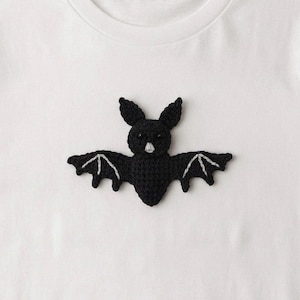 May include: A white t-shirt featuring a black crocheted bat appliqué centered on the chest. The bat has small pointed ears, a light gray stitched nose, and wings detailed with light gray thread accents that define the wing structure.