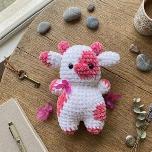May include: A handmade crochet cow plushie in white with pink spots and accents. The stuffed animal has black safety eyes and is decorated with pink flowers. The cow is sitting on a wooden surface with other decorative items.