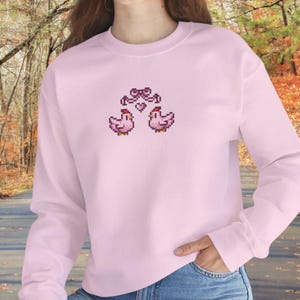 May include: A light pink sweatshirt featuring a pixelated design of two pink chickens with a bow and heart above them. The long-sleeved top is made of a soft material and is suitable for casual wear.
