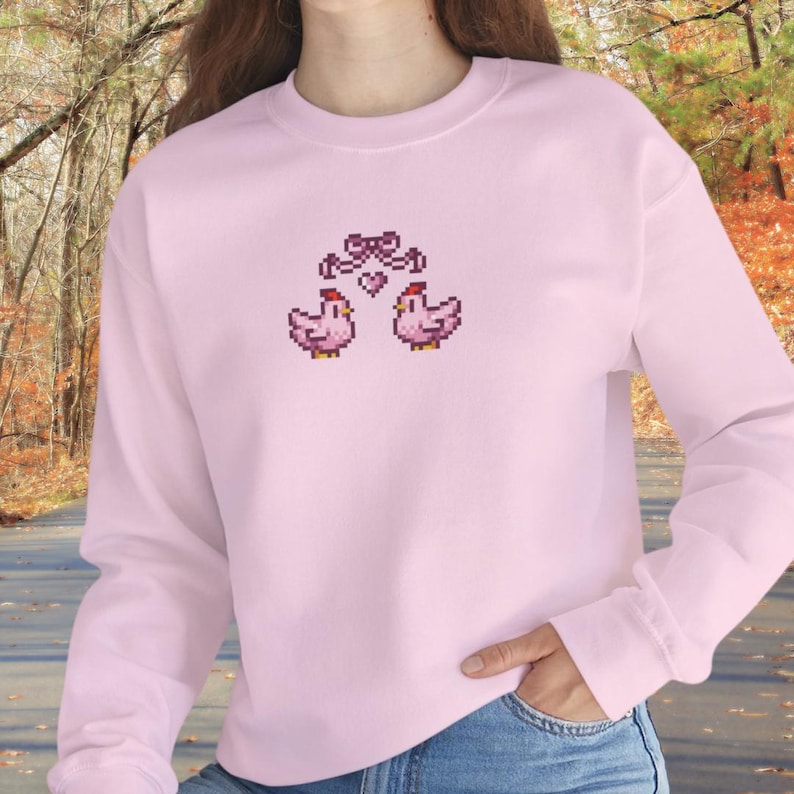 May include: A light pink sweatshirt featuring a pixelated design of two pink chickens with a bow and heart above them. The long-sleeved top is made of a soft material and is suitable for casual wear.