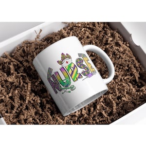 May include: White ceramic mug with the word "NURSE" in colorful, decorative lettering. The design includes a nurse's hat, stethoscope, and syringe. The mug is presented in a white box filled with brown crinkle paper.