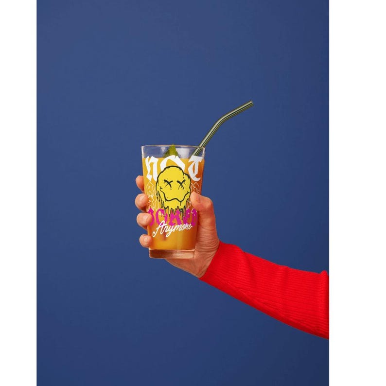 May include: A clear glass with a yellow smiley face design and the words "I'm not okay anymore" is held against a blue background. The glass contains a yellow liquid and a metal straw. The person holding the glass is wearing a red sweater.