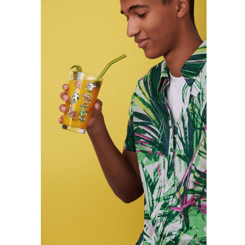 May include: A tall glass filled with orange liquid and a green straw. The glass is decorated with cartoon characters. The person is wearing a white shirt and a green and white patterned shirt. The background is yellow.