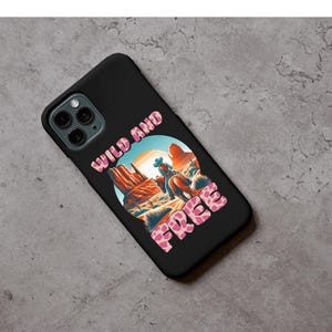 May include: Black phone case featuring a desert landscape with a cowboy on horseback. The words "WILD AND FREE" are printed in a pink, patterned font. The case has a glossy finish and a camera cutout.