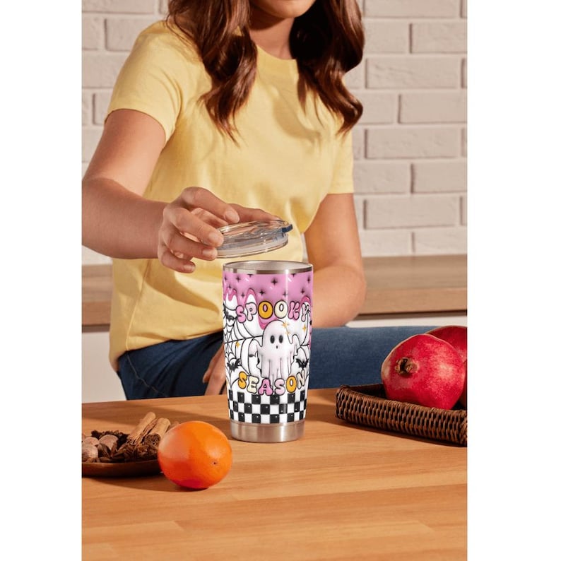 May include: A stainless steel tumbler with a pink and white Halloween-themed design. The design includes the words "Spooky Season", a ghost, spiderwebs, and a checkered pattern. The tumbler is cylindrical and has a clear lid.