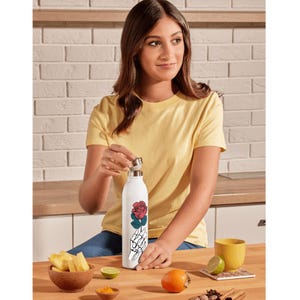 May include: White water bottle with a red rose and skeleton hand design. The bottle is being opened by a person wearing a yellow t-shirt. Other items on the table include a bowl of pineapple, a yellow mug, and a persimmon.