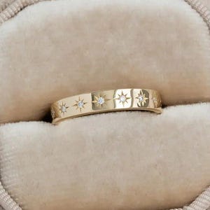 Puede incluir: Un anillo de oro con un diseño de estrella y pequeños diamantes, exhibido en un joyero de terciopelo beige. El anillo tiene un patrón repetido de estrellas y diamantes. El joyero es octogonal.