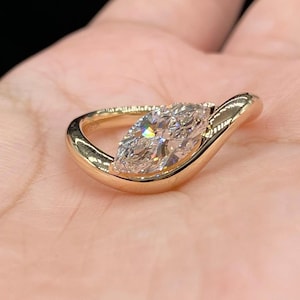May include: A marquise-cut diamond set in a gold pendant. The diamond is clear and faceted, reflecting light. The gold setting has a smooth, polished finish, with a curved design that cradles the diamond. The pendant is held in a hand.