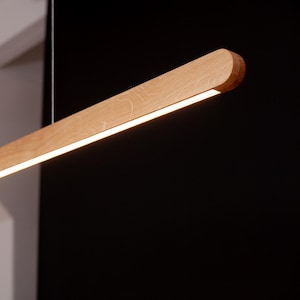 May include: A linear pendant light fitting featuring a long, horizontal wooden beam with a natural light brown wood grain finish. The underside of the beam contains a continuous integrated LED light strip that emits a warm white glow.