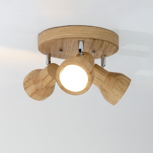 May include: A wooden ceiling light fixture with three adjustable spotlights. The fixture has a round, light-colored wood base and chrome accents. The spotlights are made of the same wood and are angled downwards, with bright white light emanating from them.