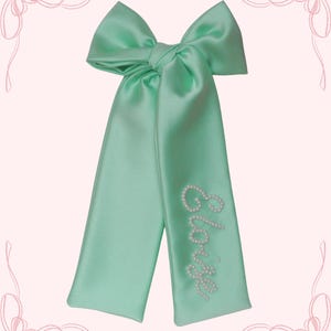 May include: A mint green satin hair bow with long streamers. The bow is tied in a large knot, and the streamers are embroidered with the name "Eloise" in white pearls. The bow is a light, pastel color.