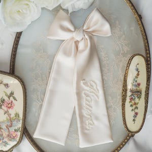 May include: A cream satin bow with long streamers, featuring the word "Yours" in pearls. The bow sits on a vintage vanity tray with a floral design, surrounded by other vintage items.