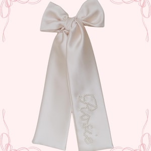 May include: A blush-colored satin hair bow with long streamers. The word "Bride" is spelled out in small, white pearls on one of the streamers. The bow is tied at the top, creating a large, decorative knot.