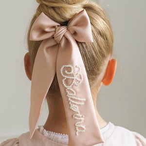 May include: A pale pink satin hair bow with long streamers, featuring the word "Ballerina" in white pearls. The bow is styled in a bun.
